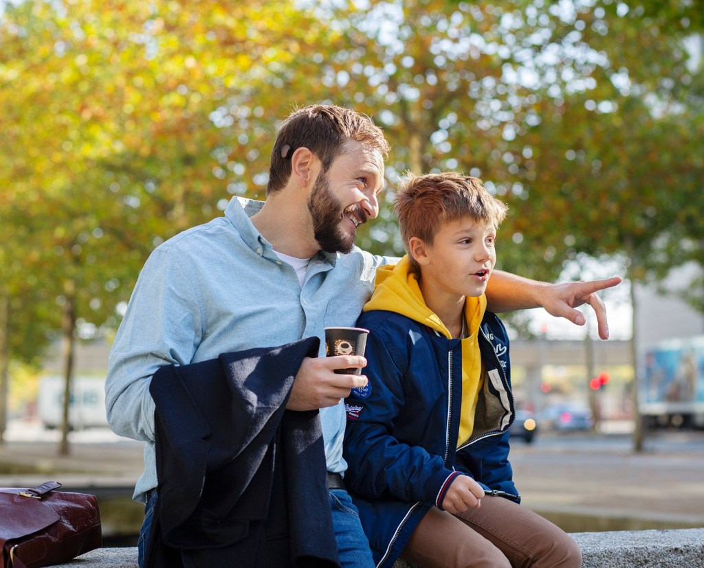 Father and son sitting on a wall with trees behind them. The father has a bone-anchored hearing aid (BAHA). He is holding a cup of coffee and pointing to in the direction that the child is looking.