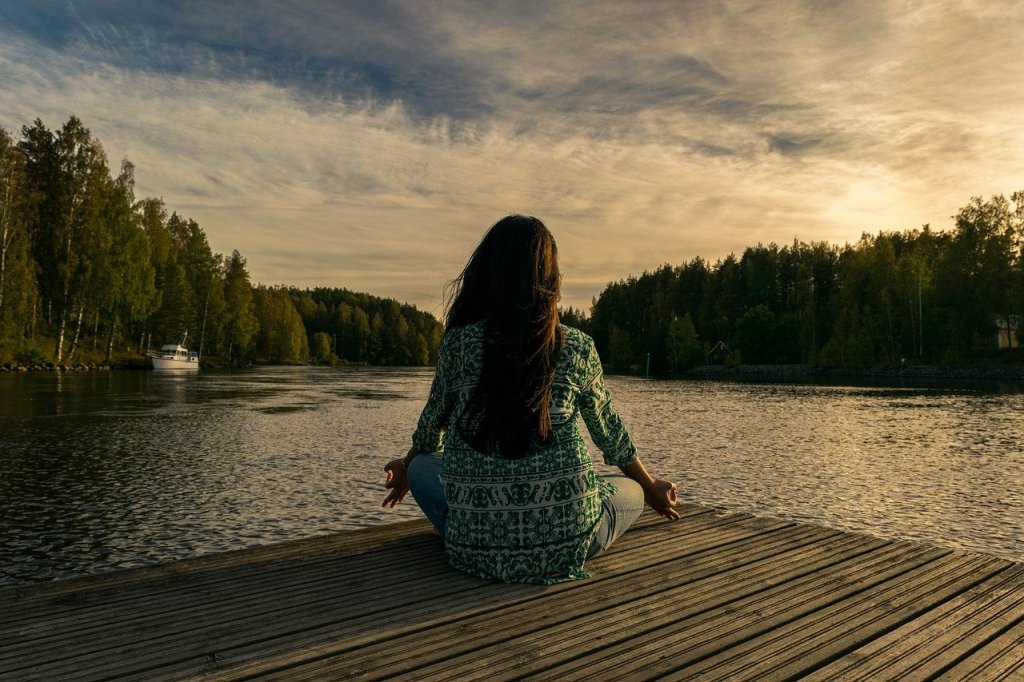 Woman sitting on decking by water in meditation pose. 