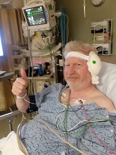 Shai in hospital after cochlear implant surgery with a dressing over his left ear. He is smiling and giving a 'thumbs up.'