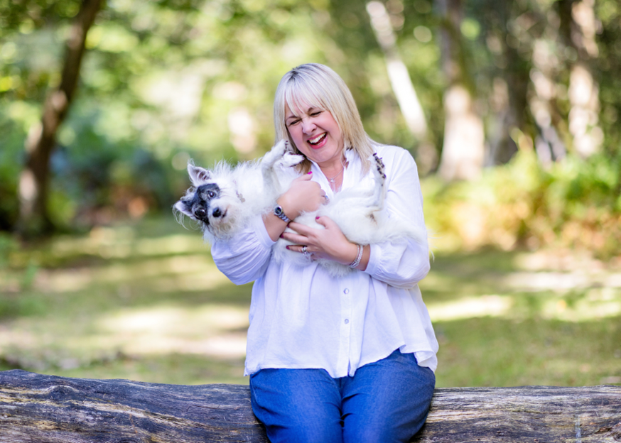 Blond haired woman, sitting on a tree trunk and holding her fluffy black and white dog. They are both smiling.